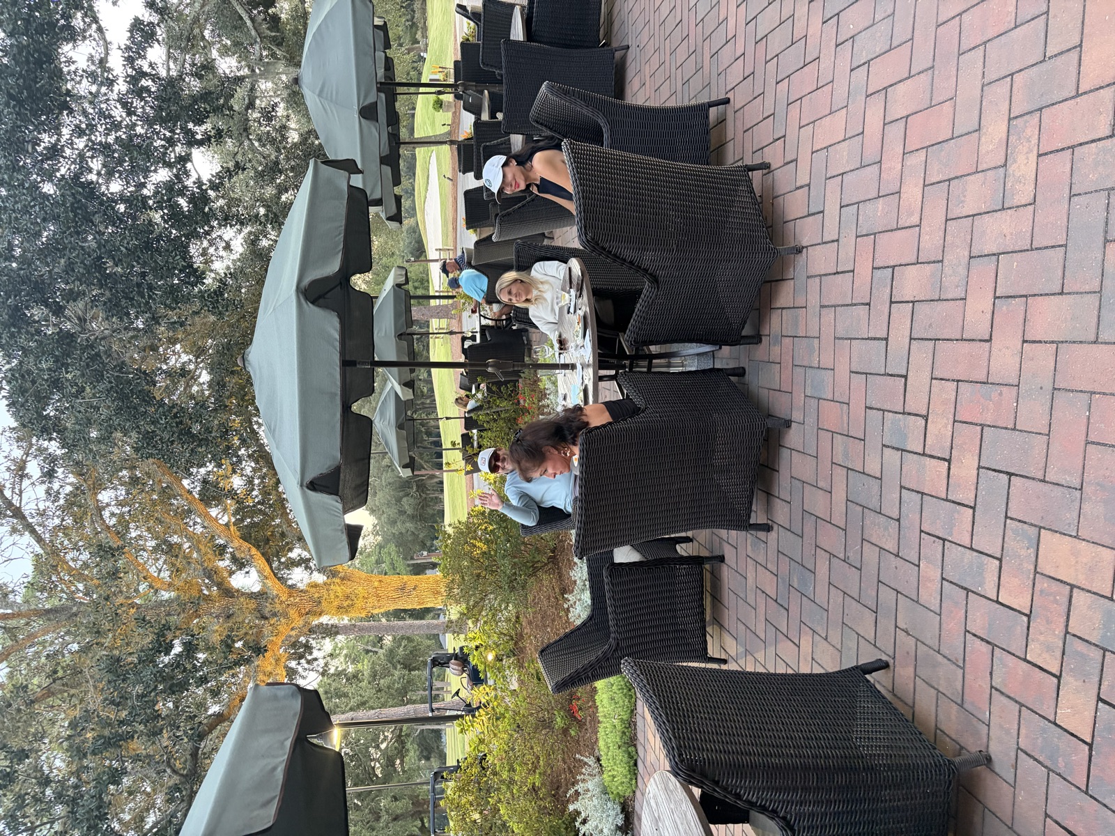 Four people at a patio table under an umbrella, one waving at the camera